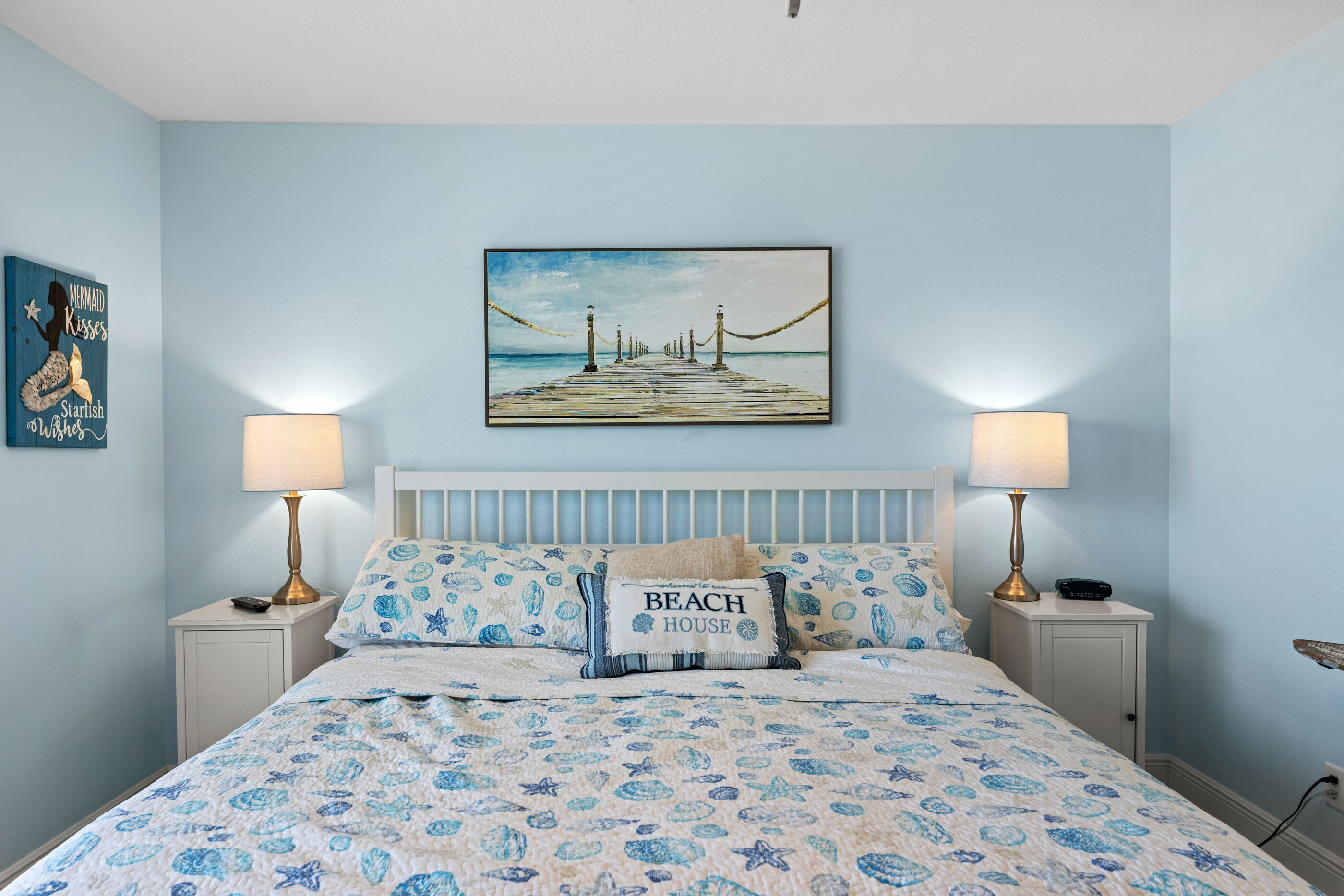 A bedroom with a bed, two bedside tables, a nightstand, a headboard, a painting of a pier, and a wall decoration.