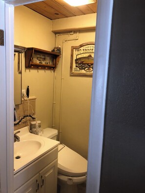 Et lille badeværelse med en hvid vask, toilet og spejl. Der er en træhylde over spejlet og et velkomstskilt i ramme på væggen.
