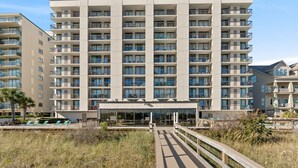 A multi-story hotel with a central entrance, balconies on each floor, and a pool area visible in the background.