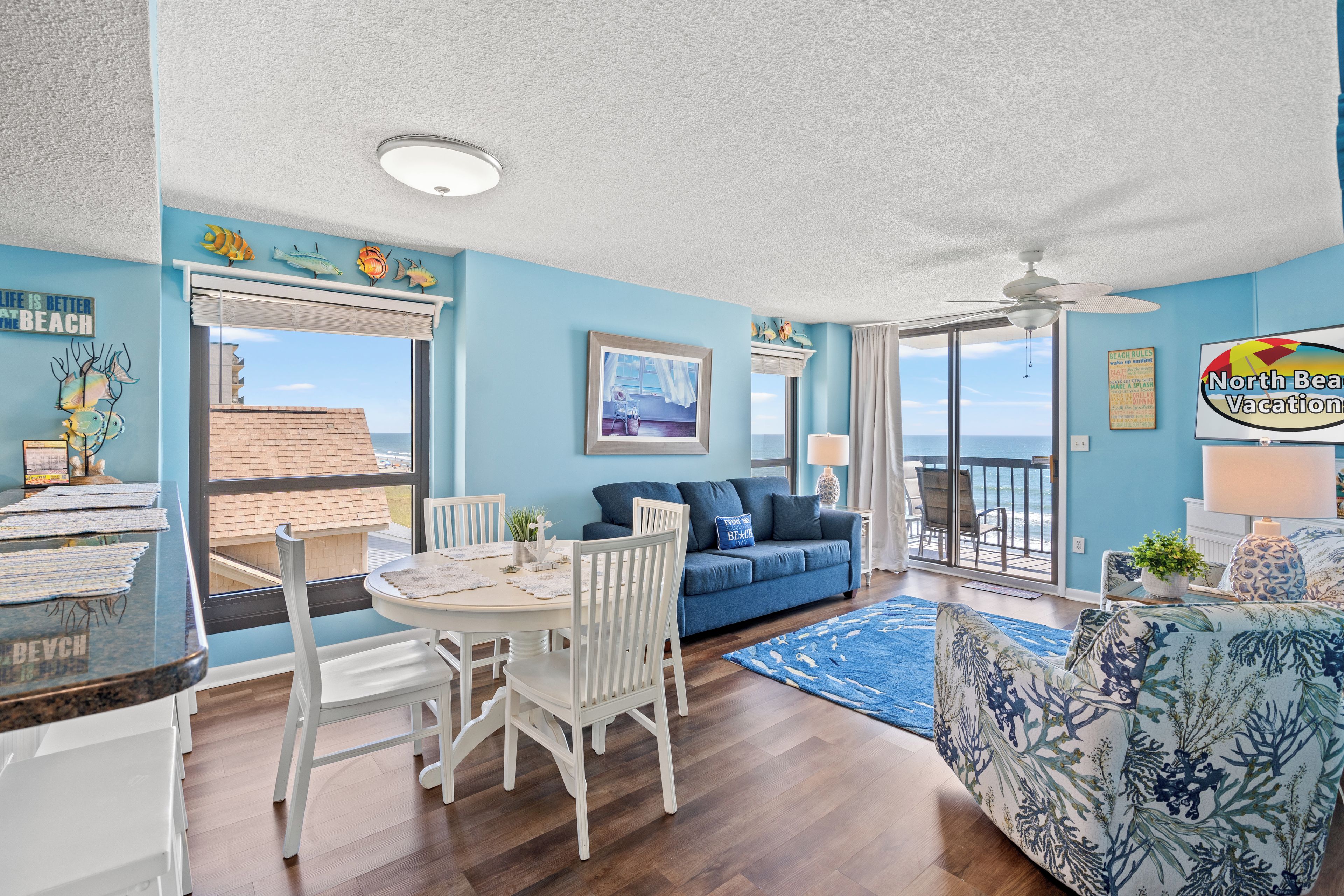 A room with a dining table, chairs, a sofa, a balcony with a view of the sea, and a ceiling fan.