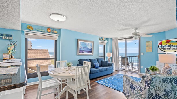 A room with a dining area, a blue sofa, and a view of the beach.