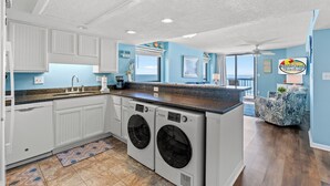 A laundry room with two front-loading washers and dryers, a countertop with a coffee maker, and a view of the ocean through large windows.