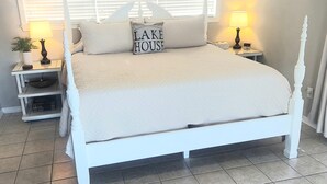 A white four-poster bed with a 'LAKE HOUSE' pillow, two bedside lamps, a small plant, and a nightstand with a clock.