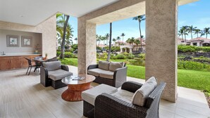 A spacious outdoor area with wicker furniture, a round coffee table, and a view of a well-maintained garden and buildings in the background. - 🏖️Upgraded Waikoloa Condo w/ Central A/C & Lanai (Waikoloa)