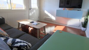 A living room with a sofa, a table, a television mounted on the wall, and a potted plant.