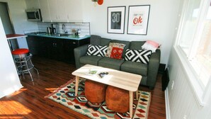 A modern living room with a sofa, coffee table, and patterned rug. A kitchen with a microwave and cabinets is visible in the background.