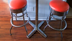 A red table with two stools, a framed sign above it, and a wooden floor.