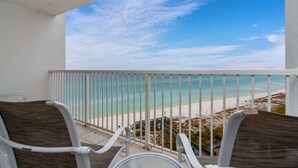 Ein Balkon mit zwei Stühlen und einem Tisch, der auf einen Strand mit blauem Himmel blickt.