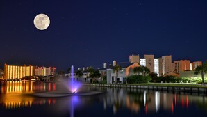 Ein nächtliches Stadtbild bei Vollmond, beleuchtete Gebäude und eine Fontäne, die sich im Wasser spiegelt.