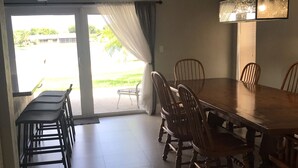 Et spiseområde med et stort træbord og stole, en moderne pendellampe samt udsigt gennem en glasdør til et udendørs område.