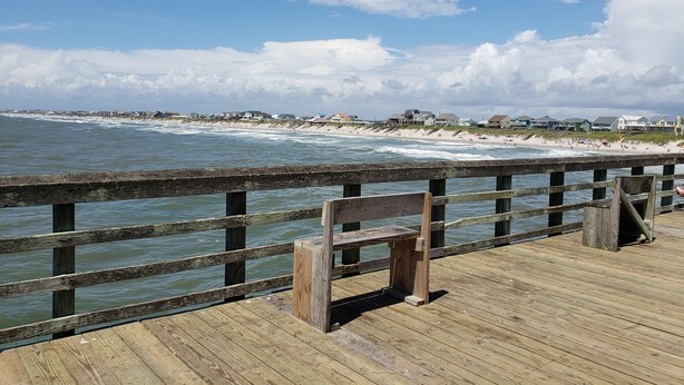 Un muelle de madera con un banco, con vista a una playa con casas y el océano.
