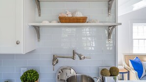 A kitchen with white tiled backsplash, stainless steel fixtures, and a basket of decorative items on a shelf. - New In Town Time & Tide Cottage (Nantucket)