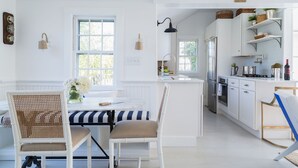 A bright kitchen with white cabinetry, a dining area with striped tablecloths, and a view of the outdoors. - New In Town Time & Tide Cottage (Nantucket)