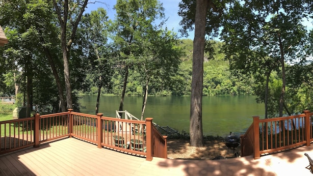 Una terraza con vista a un lago rodeado de árboles, una parrilla para barbacoa y mobiliario de exterior.