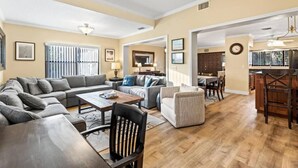 A spacious living room with a large sectional sofa, a coffee table, and a dining area with a kitchen in the background. - TIKI BLUE SOUTH: Walk to Beach + Heated Pool (Satellite Beach)