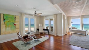 Ein geräumiges Wohnzimmer mit Meerblick, Holzboden, Deckenventilator und einem großen abstrakten Gemälde.