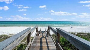 Ein hölzerner Steg führt zu einem Strand mit klarem türkisblauem Wasser und sandigem Ufer.