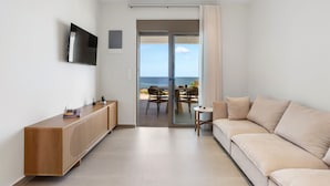 A modern living room with a sofa, TV, wooden cabinet, and a sliding glass door leading to a balcony with a view of the sea. - Seafront Junior Villa with private heated Jacuzzi (Chania)