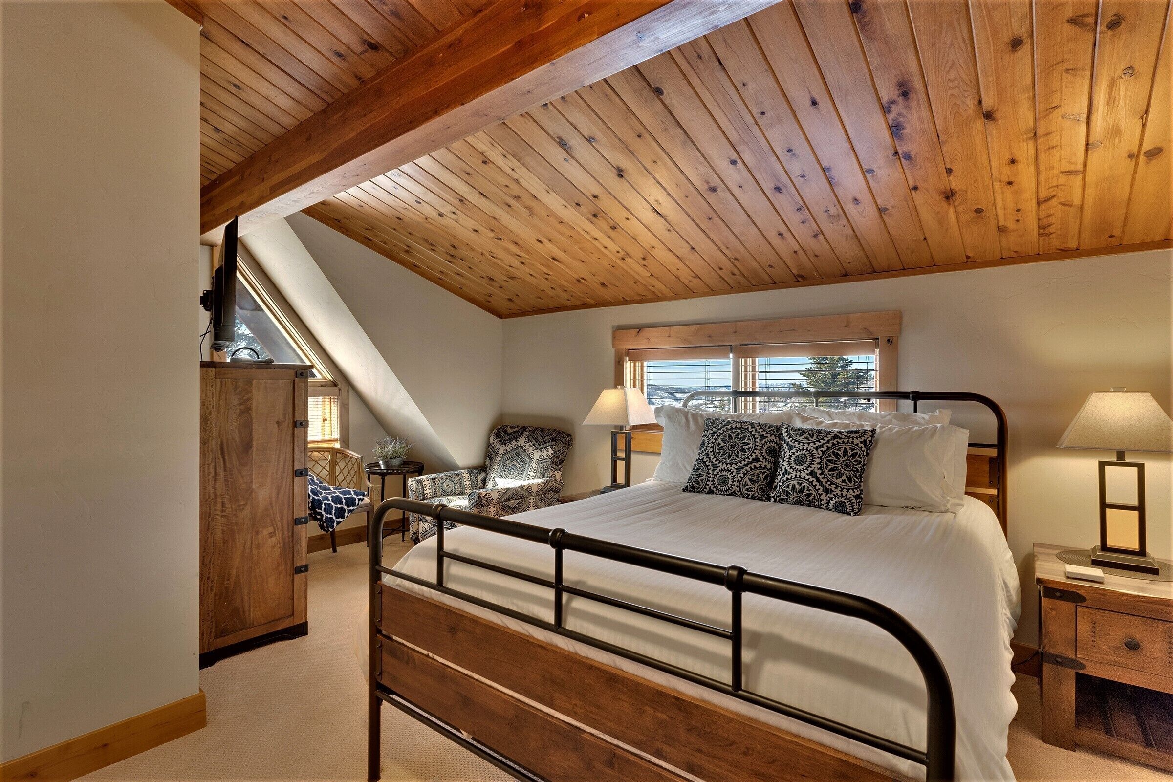 A bedroom with a wooden ceiling, a bed with a metal frame, a nightstand, a lamp, and a window with blinds.