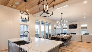 A modern kitchen with a large island, a dining area with a chandelier, and a living room with a TV. - Coastal Manor~ 6 bdrm, across from ocean,  Ocean Views!, Private Pool and Hot Tub, Elevator! (Isle Of Palms)