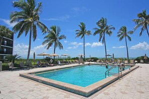 A swimming pool with lounge chairs and umbrellas, surrounded by palm trees. - Sundial B306--Direct Beachview, Newly Renovated, Steps to Beach (Sanibel Island)