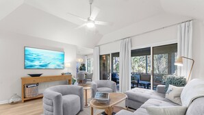 A modern living room with a ceiling fan, a large TV, grey sofas, a wooden coffee table, and a view of the outdoors through large windows. - Villa Palma, newly renovated, central, in popular location (Johns Island)