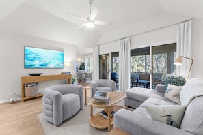 A modern living room with a ceiling fan, a large TV, grey sofas, a wooden coffee table, and a view of the outdoors through large windows.