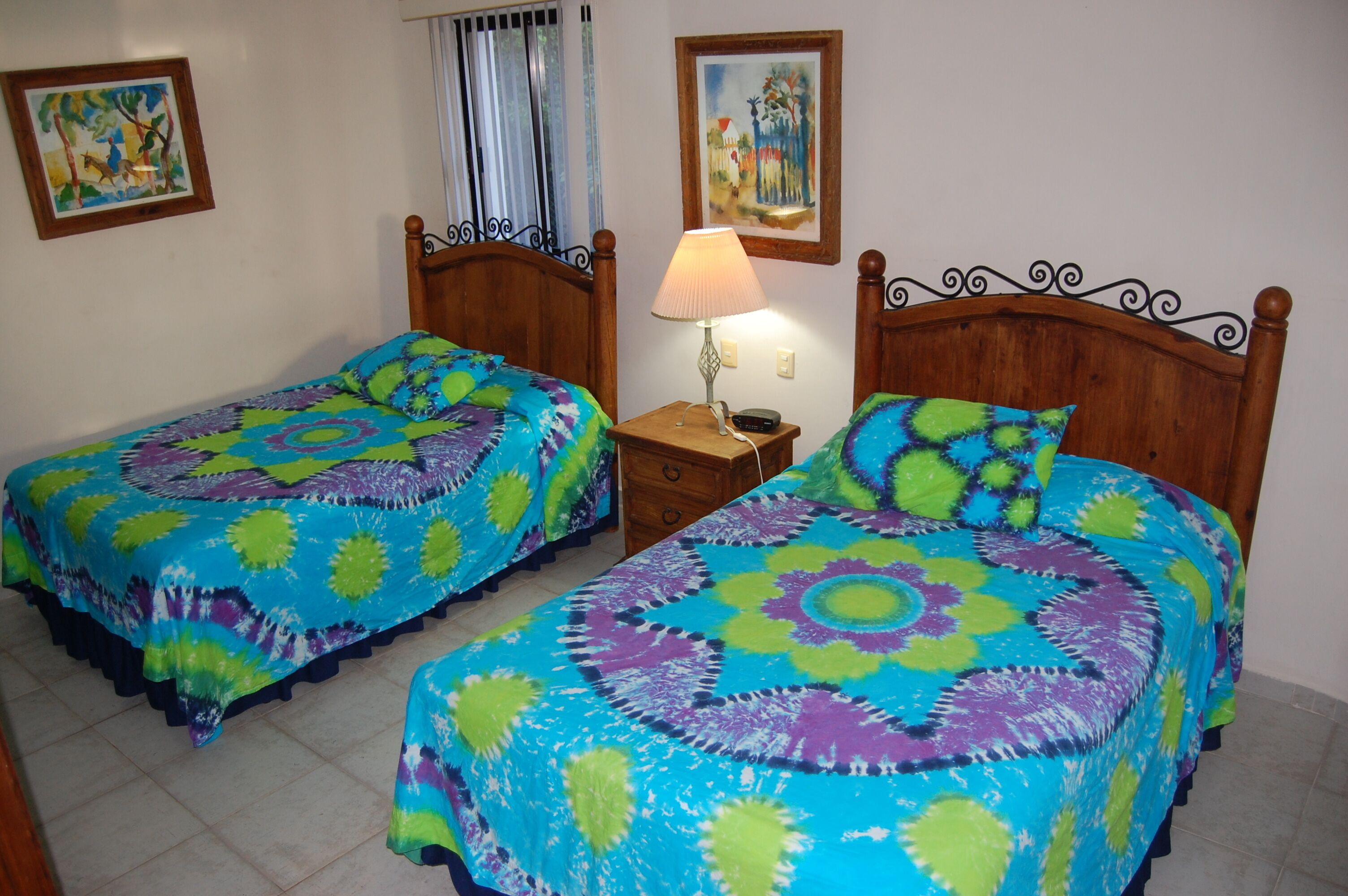 Dos camas individuales con ropa de cama colorida, una mesita de noche con lámpara y dos cuadros en la pared.