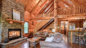 L’intérieur d’une cabane en rondins, avec une cheminée en pierre, un escalier en bois et un salon confortable comprenant un canapé et une table basse.