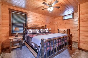 Ein Schlafzimmer einer Holzhütte mit einem Bett, zwei Nachttischen, einem Deckenventilator und einem Fenster mit Jalousien.