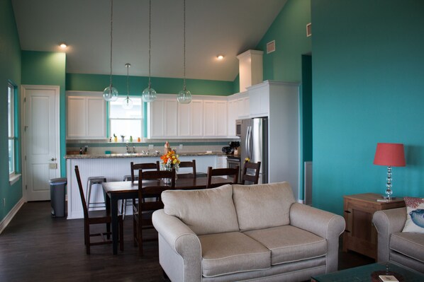 Una cocina moderna con armarios blancos, una zona de comedor con sofá color beige y paredes azul verdoso.