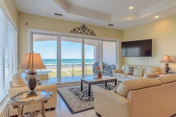 A living room with a sofa, a coffee table, and a TV. There is a large window with a view of the beach.