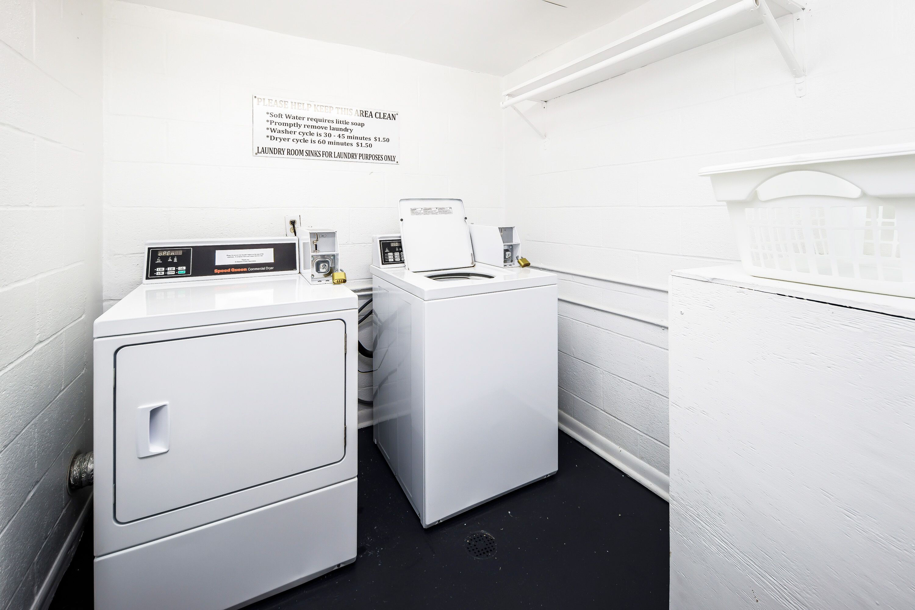 A laundry room with a washing machine, dryer, and stacked laundry baskets. A sign instructs to keep the area clean and lists laundry-related rules.