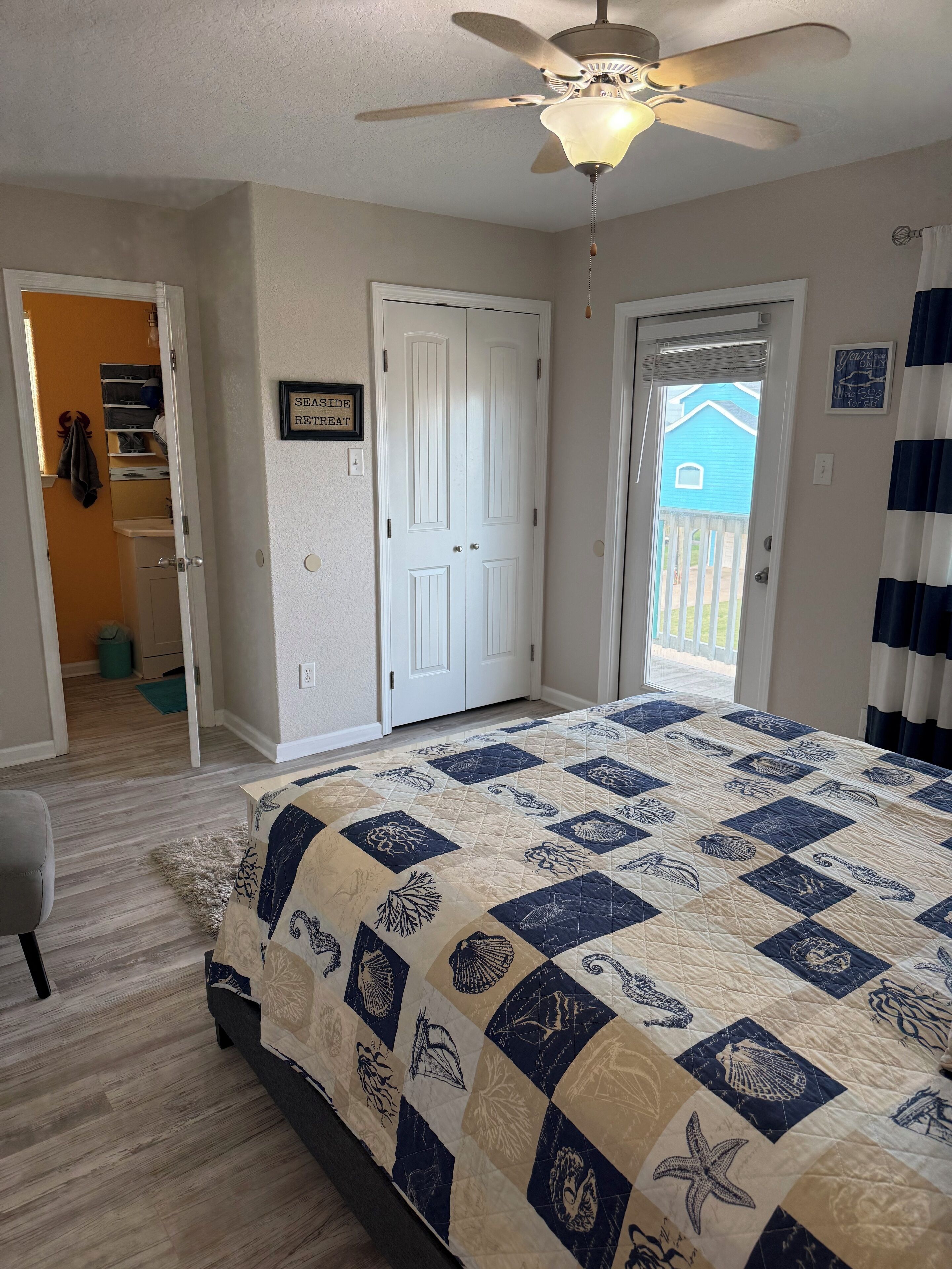 A bedroom with a bed, a ceiling fan, a door, and a view of the outside.
