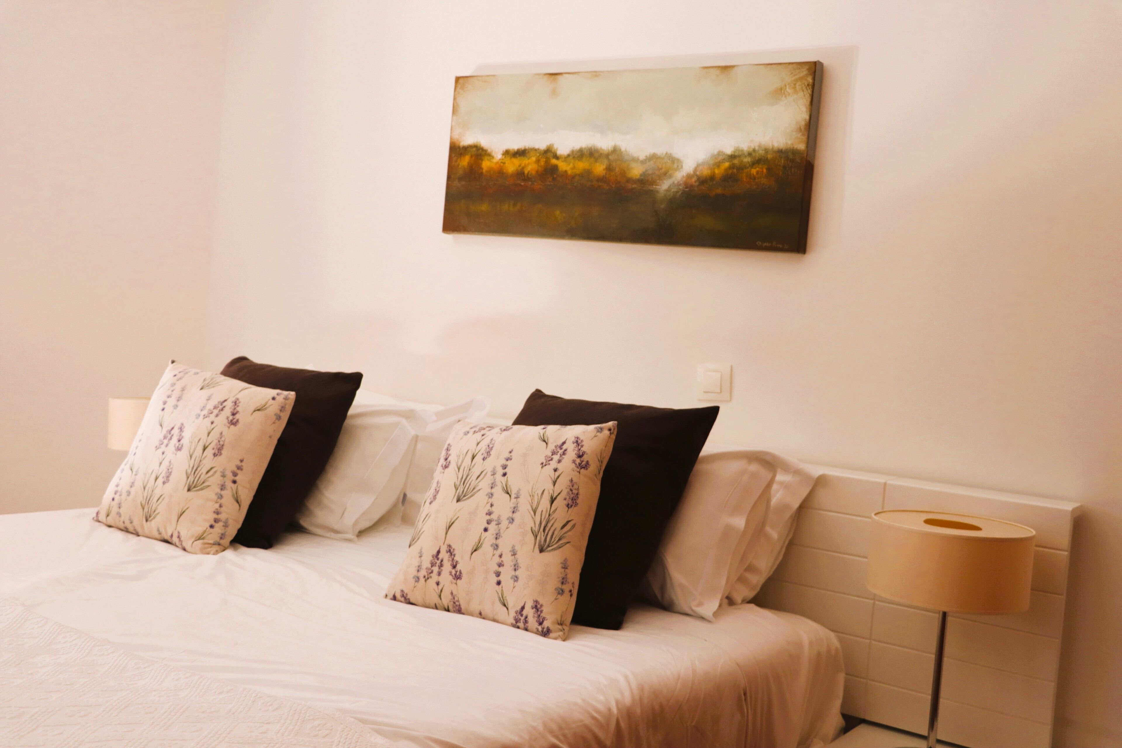 A bedroom with a bed, pillows, a bedside lamp, and a painting on the wall.