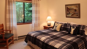 Um quarto com uma cama, uma cadeira, uma mesinha de cabeceira com abajur, uma janela com cortinas e um quadro na parede.