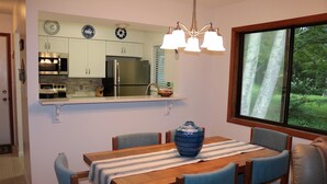 Uma sala de jantar com uma mesa de madeira, cadeiras azuis e um vaso azul. Uma cozinha é visível ao fundo, com armários brancos e eletrodomésticos de aço inoxidável.