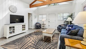 A spacious living room with a high ceiling, wooden beams, a flat-screen TV, a white cabinet, a patterned rug, a blue sofa, and a coffee table. - Pup Friendly Home w/ Fully Fenced Yard! (Hilton Head Island)