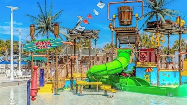 Ein Wasserpark mit einer grünen Rutsche, einem Spritzbereich und einer hölzernen Spielkonstruktion.