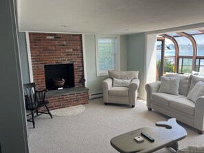 Una sala de estar con chimenea de ladrillo, un sillón blanco con un cojín, un sofá color crema y vistas al agua a través de amplias ventanas.