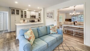 A spacious living room with a blue sofa, a kitchen with white cabinets, and a dining area with a chandelier. - Pet Friendly 3BR w/ Screened Porch & Amenities (Hilton Head Island)