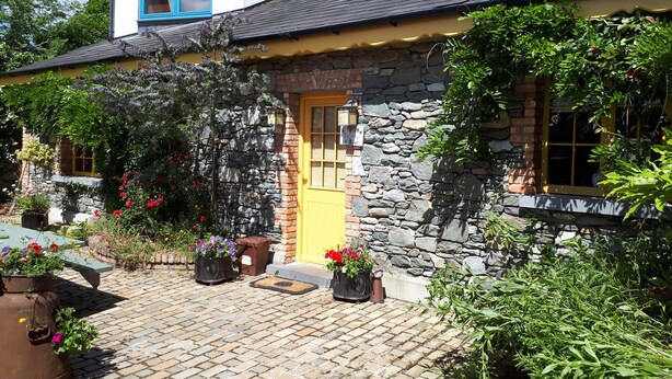 Une maison en pierre avec une porte jaune, entourée de plantes en pot et dotée d’une terrasse pavée.