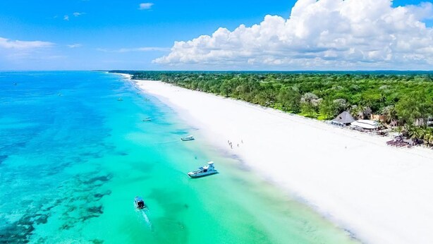 Ein Strand mit klarem türkisblauem Wasser, weißem Sand und einer Baum- und Gebäudezeile entlang der Küste.