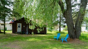Una pequeña cabaña de madera con un porche, dos sillas azules y un árbol grande al frente.