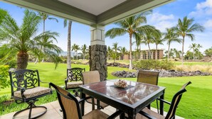 A covered outdoor seating area with a table and chairs, surrounded by a lush garden with palm trees and a view of a building in the background.