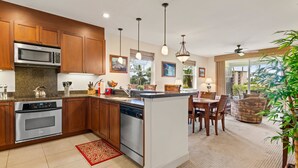 A modern kitchen with wooden cabinets, stainless steel appliances, and a central island. There is a dining area with a table and chairs, and a view of the outdoors through large windows.