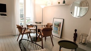 Ein modernes Wohnzimmer mit Glastisch, Holzsesseln, einem schwarzen Drahtstuhl, einem kleinen runden Tisch, einem gerahmten Bild, einem runden Spiegel und einer kleinen Statue.