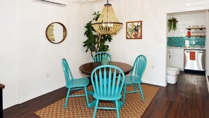 A dining area with a round table, four turquoise chairs, a woven rug, a wooden floor, a pendant light, a mirror, and a kitchen in the background. - Comfy Beach Studio for 2 - Only a short walk to the beach. (Cape Canaveral)