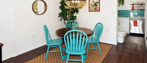 A dining area with a round table, four turquoise chairs, a woven rug, a wooden floor, a pendant light, a mirror, and a kitchen in the background.