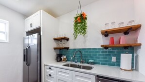 A kitchen with white cabinets, a stainless steel refrigerator, a sink, and a dishwasher. There are shelves with glassware and a hanging plant. - Comfy Beach Studio for 2 - Only a short walk to the beach. (Cape Canaveral)